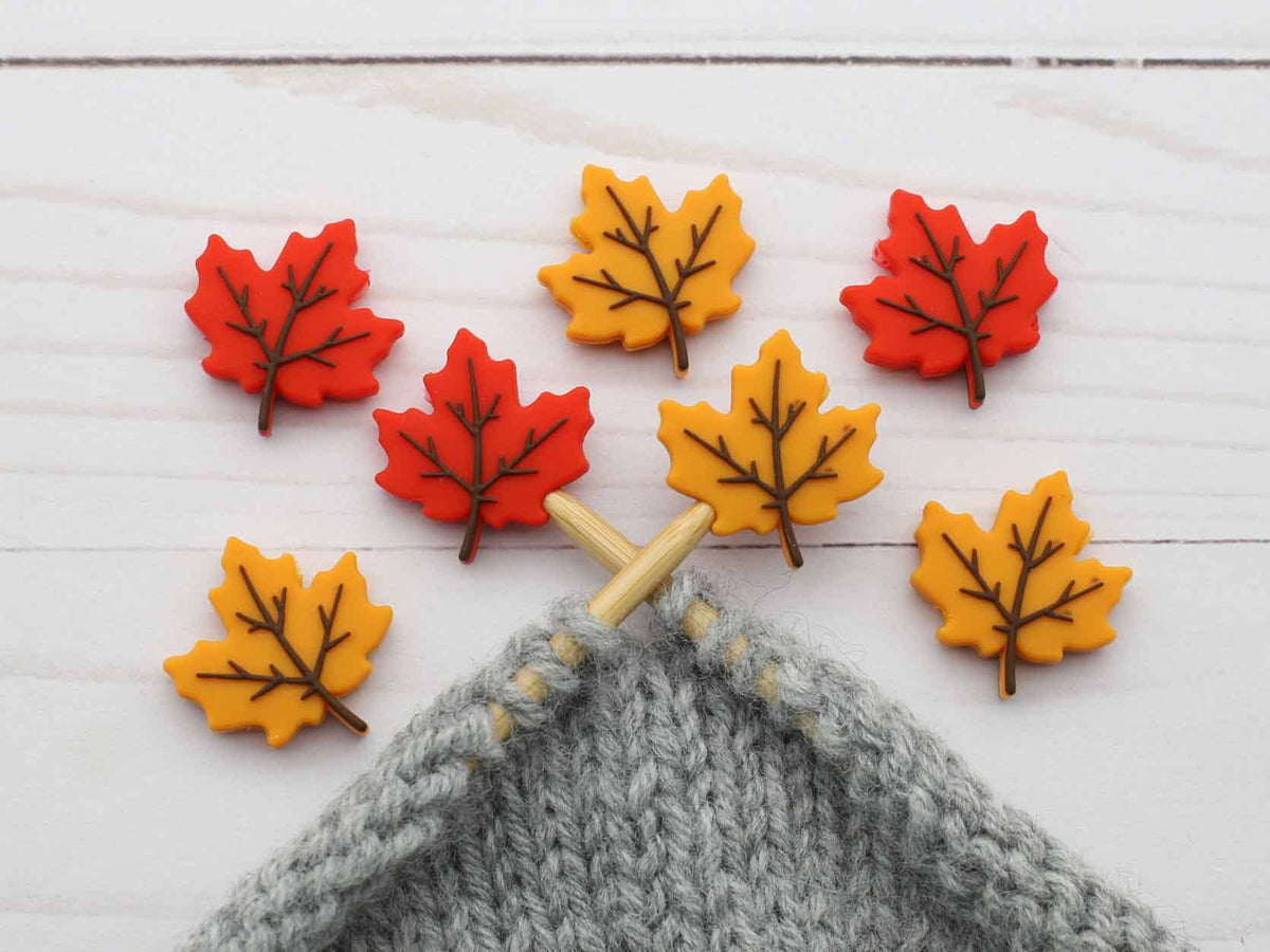Seven autumn maple leaf-shaped stitch stoppers in red, orange, and yellow with brown leaf veining, arranged on a white wood background above a gray knitted swatch with wooden knitting needles.