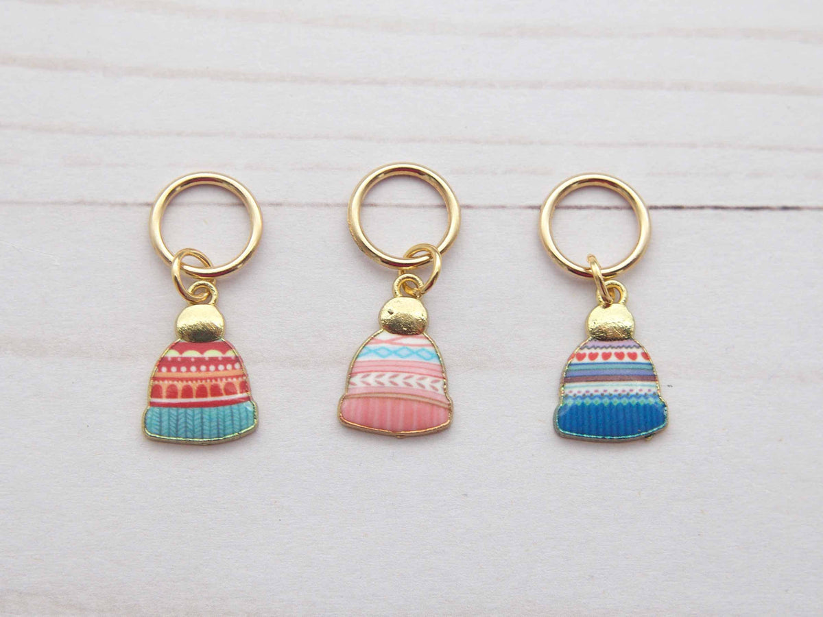 Three colorful stitch markers with gold rings on a light wooden surface