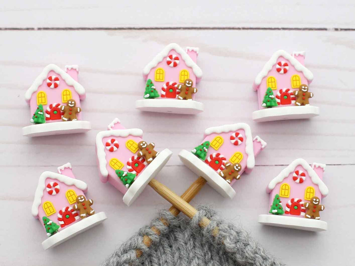 Six small decorative house-shaped stitch stoppers with pink roofs, white trim, candy motifs, and gingerbread figures, arranged on a white wood surface with knitting needles and gray yarn visible at the bottom.