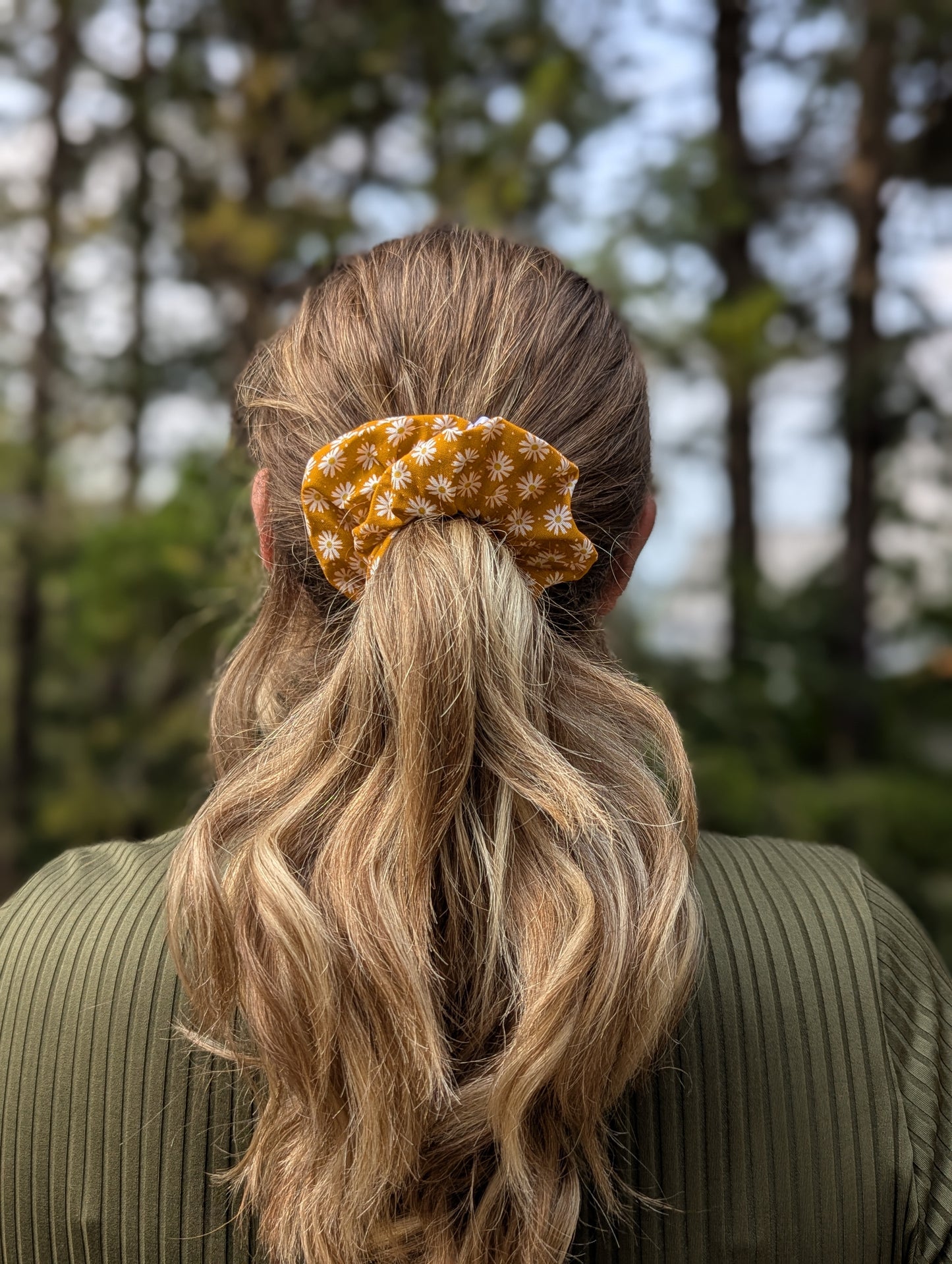Person with long blonde hair tied back with a yellow scrunchie, standing outdoors with trees in the background.