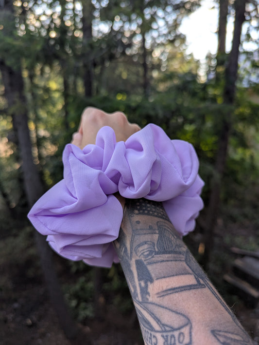 Purple scrunchies held by a hand with tattoos against a forest background