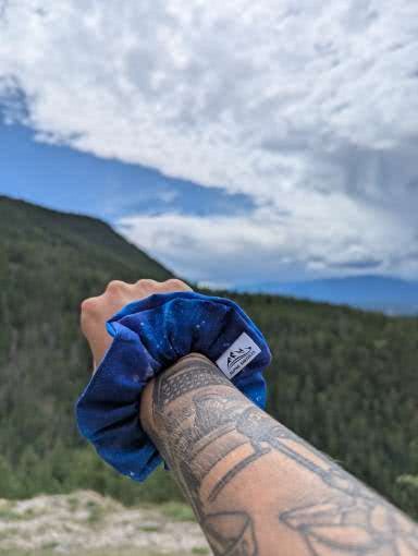 Blue scrunchie held by a tattooed arm with a mountainous landscape in the background