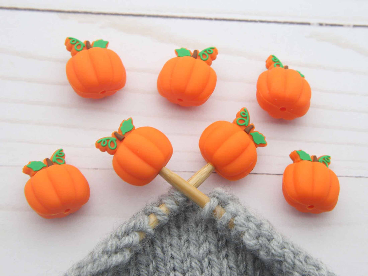 Seven small orange pumpkin-shaped Stitch stoppers with green leaves are arranged on a white wooden surface, with two on wooden knitting needles over a gray knitted fabric.