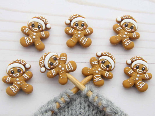 Six small gingerbread person stitch stoppers with white icing details and Santa hats are arranged on a white wooden surface, with a gray knitted yarn and wooden knitting needles visible at the bottom.