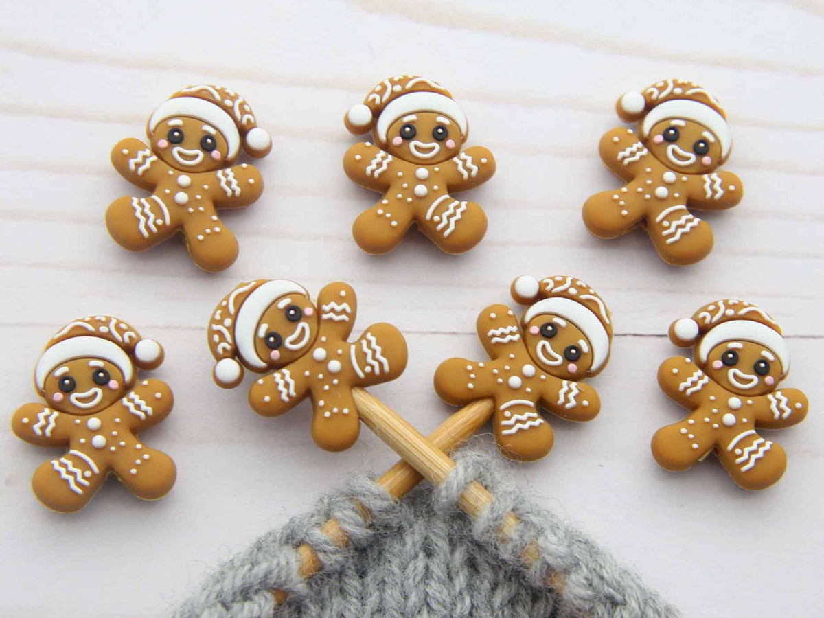 Six small gingerbread person stitch stoppers with white icing details and Santa hats are arranged on a white wooden surface, with a gray knitted yarn and wooden knitting needles visible at the bottom.