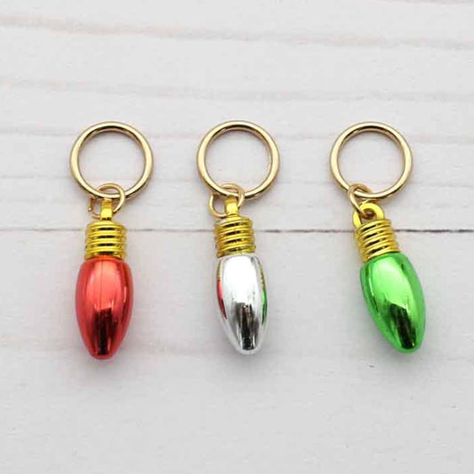 Three small Christmas light-shaped stitch markers in red, silver, and green, each with a gold-tone ring and wrapped top, arranged on a white wood background.