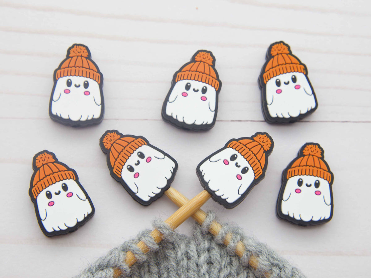 Six cute ghost-shaped stitch stoppers on knitting needles with orange knit hats and pink cheeks, arranged on a white wooden background with a gray knitted piece at the bottom.