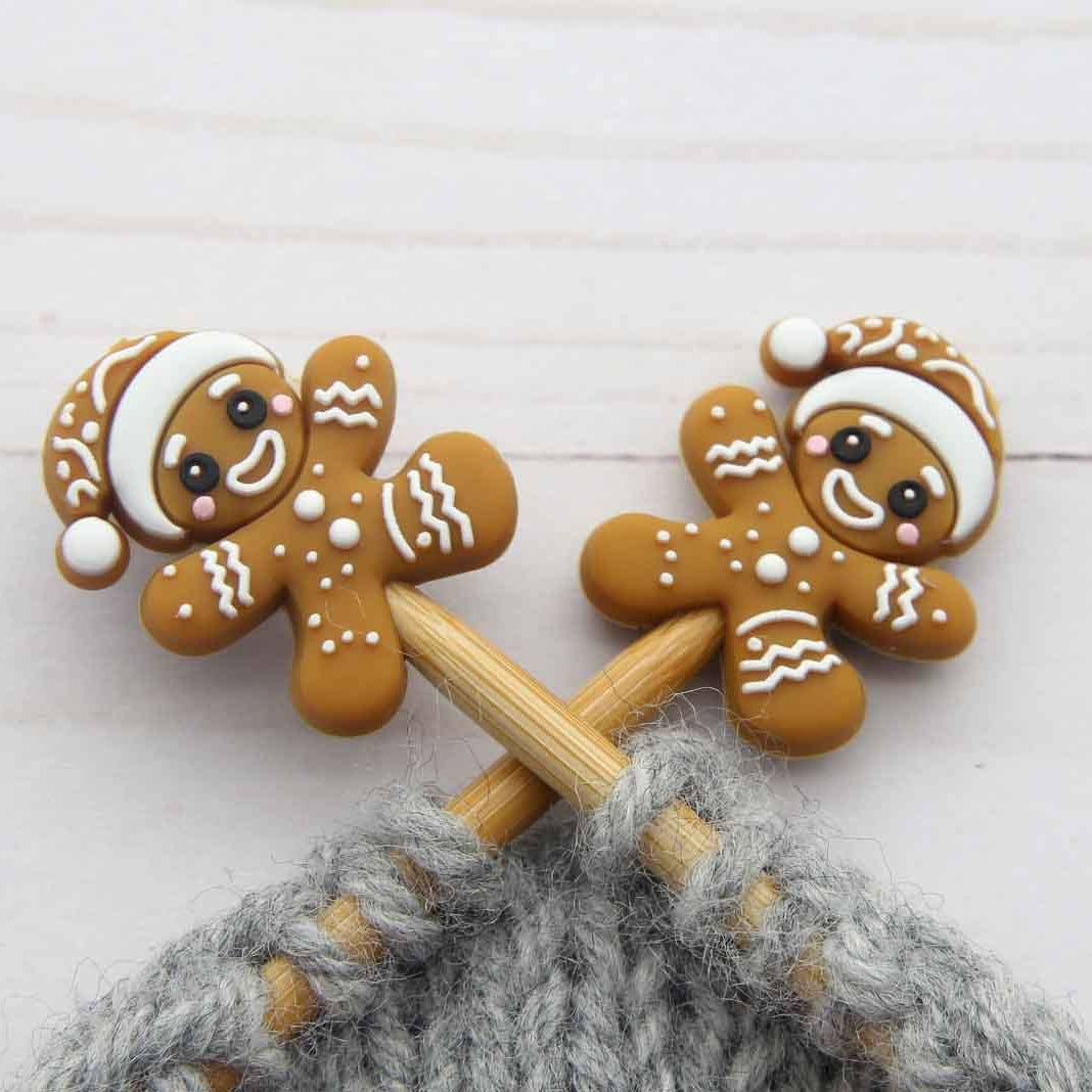 Two gingerbread person-shaped stitch stoppers on knitting needles, decorated with white icing-like patterns and Santa hats, on a white wooden surface.