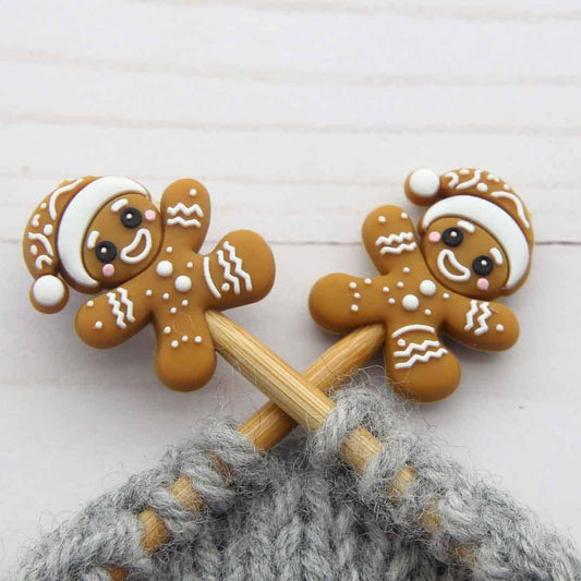 Two gingerbread person-shaped stitch stoppers on knitting needles, decorated with white icing-like patterns and Santa hats, on a white wooden surface.
