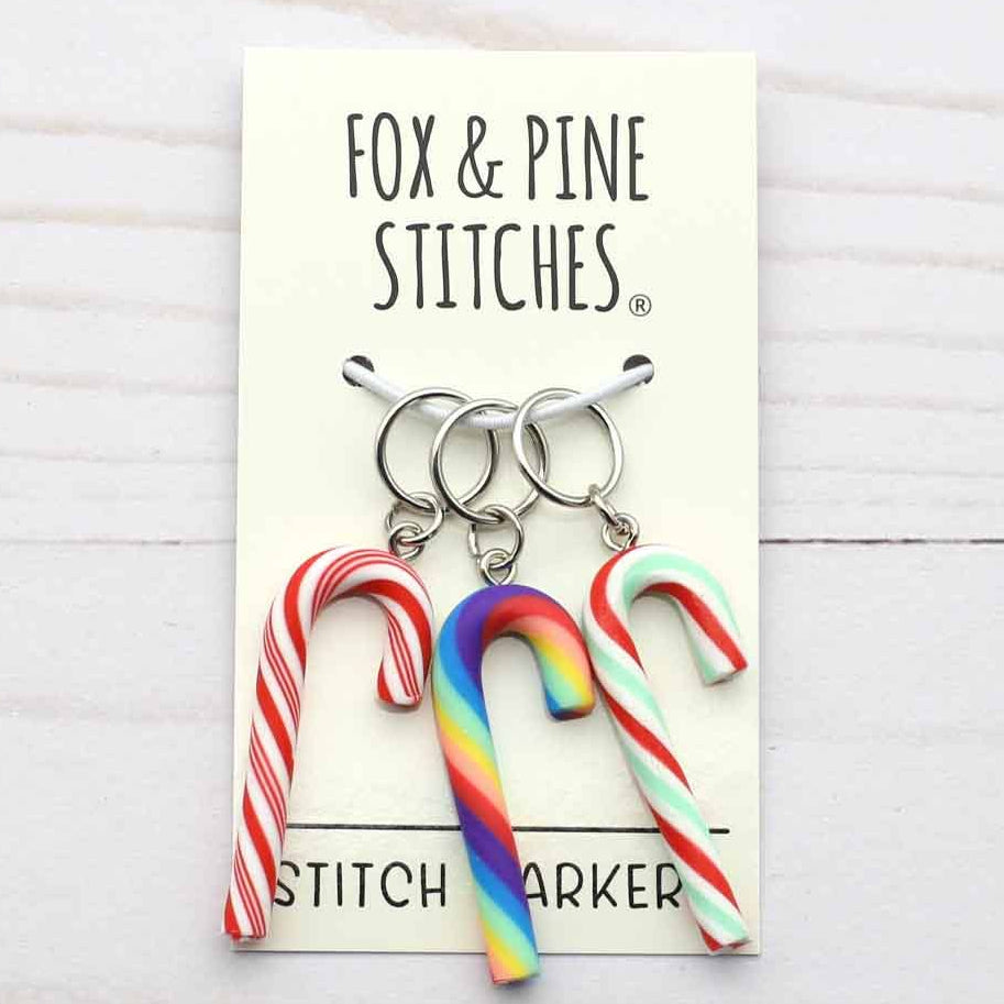 Three candy cane-shaped stitch markers with silver rings attached to a cream display card, branded 'FOX & PINE STITCHES', set against a white wood background.