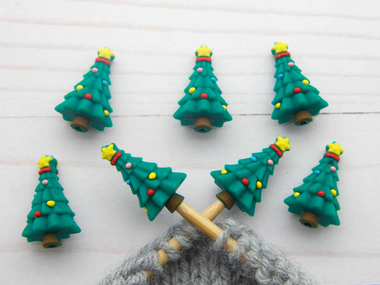 Six small Christmas tree-shaped knitting needle stitch stoppers are arranged on a white wood background, with a gray knitted loop attached at the bottom.
