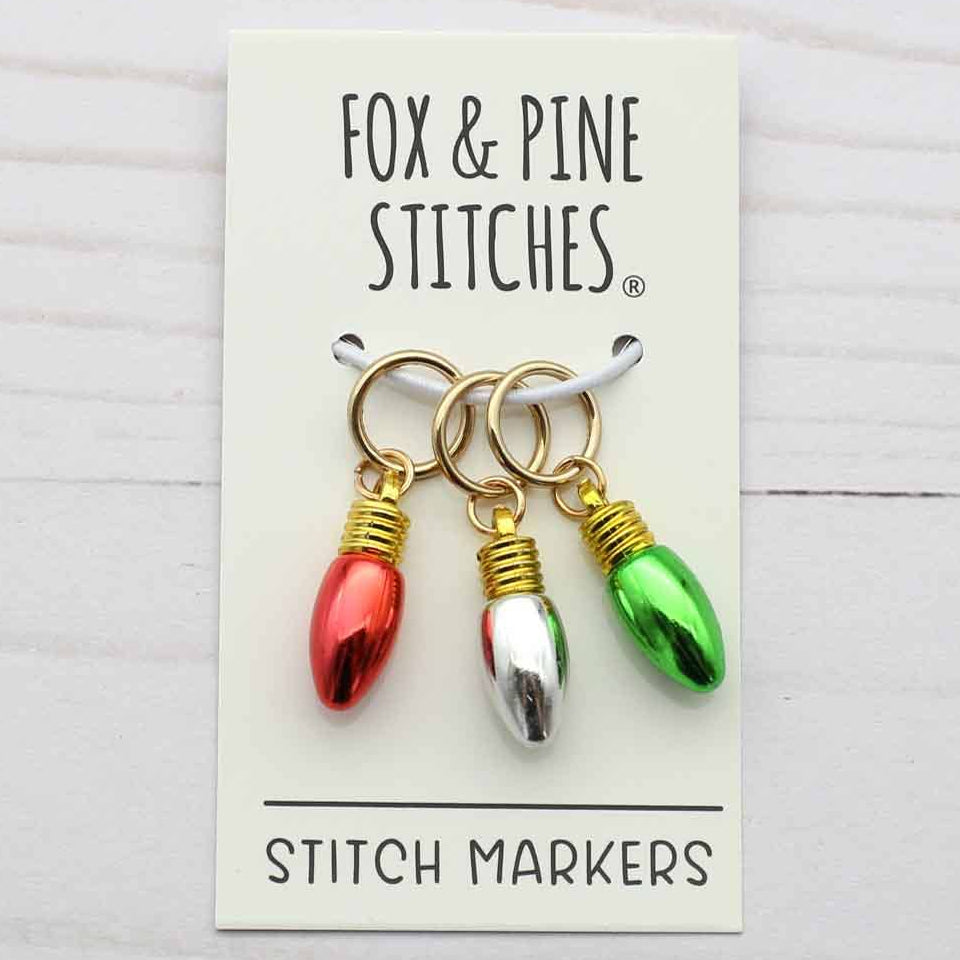 Three Christmas light-shaped stitch markers by Fox & Pine Stitches, displayed on their cream-colored backing card against a white wood background.