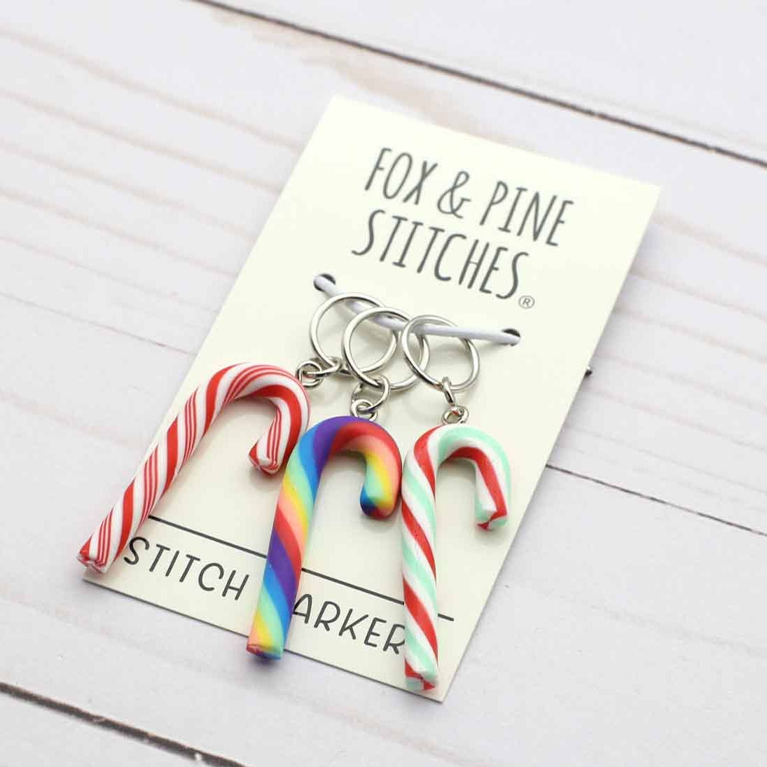 Three candy cane-shaped stitch markers by Fox & Pine Stitches, displayed on their cream-colored backing card over a white wood plank background.