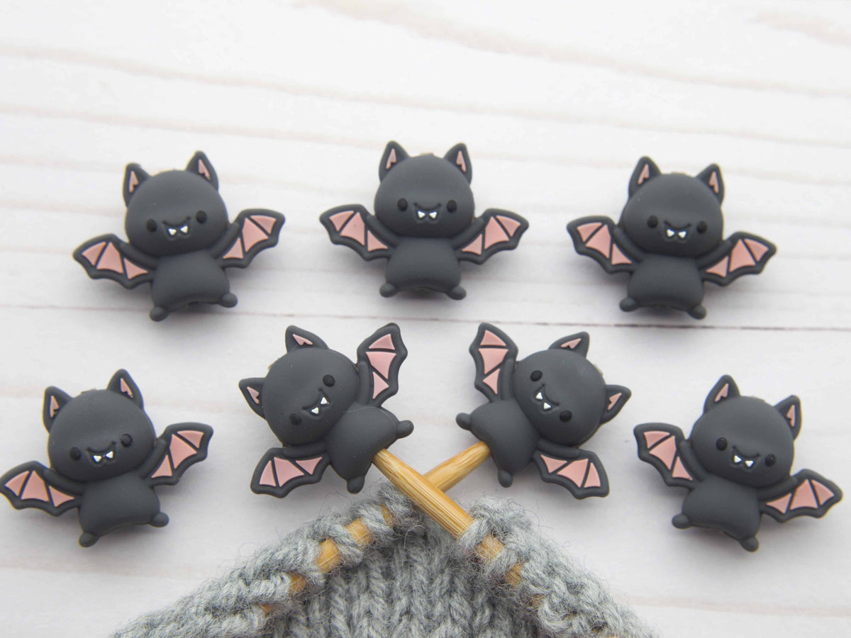 Set of cute bat-shaped knitting stitch stoppers on a wooden needle with a textured fabric background.