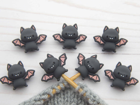 Set of cute bat-shaped knitting stitch stoppers on a wooden needle with a textured fabric background.