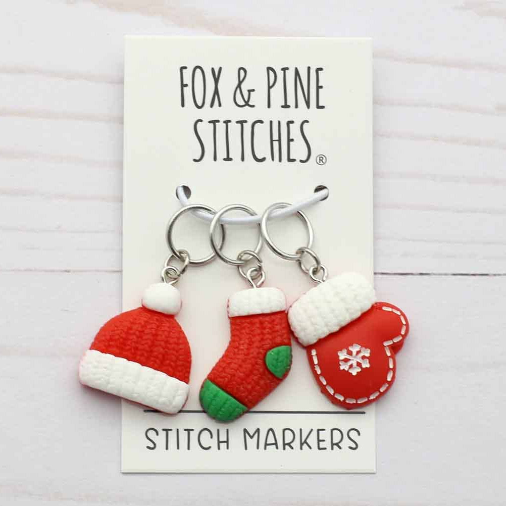 Three Christmas-themed stitch markers by Fox & Pine Stitches, featuring a Santa hat, a stocking, and a mitten on silver rings, displayed on a white card over a light wood background.