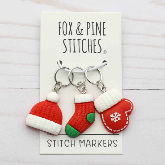 Three Christmas-themed stitch markers by Fox & Pine Stitches, featuring a Santa hat, a stocking, and a mitten on silver rings, displayed on a white card over a light wood background.