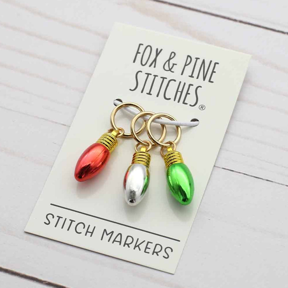 Three Christmas light-shaped stitch markers in red, silver, and green on a cream card labeled 'FOX & PINE STITCHES STITCH MARKERS' on a white wood background.