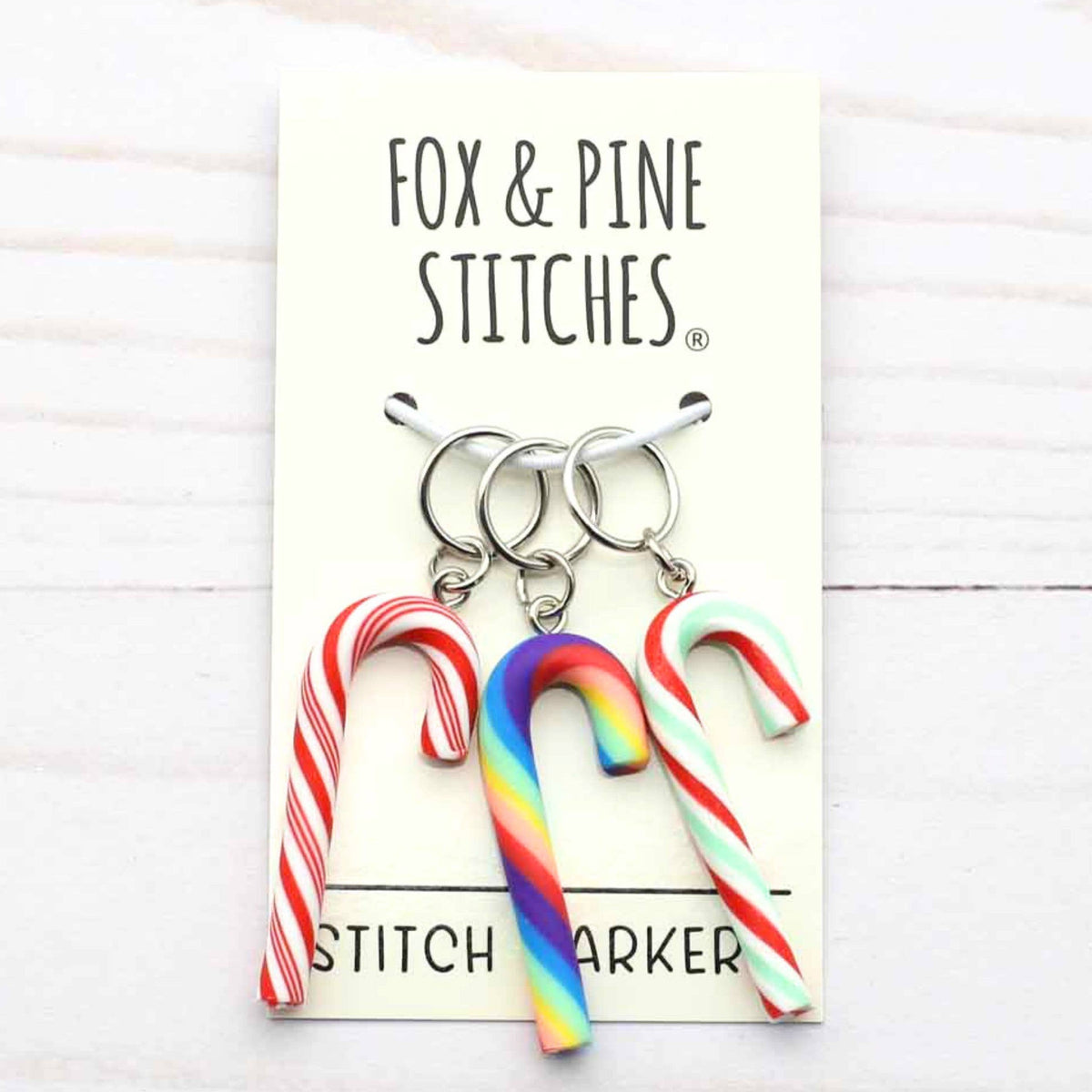 Three candy cane-shaped stitch markers with red, white, and rainbow stripes hang from silver rings on a cream display card labeled 'FOX & PINE STITCHES'.