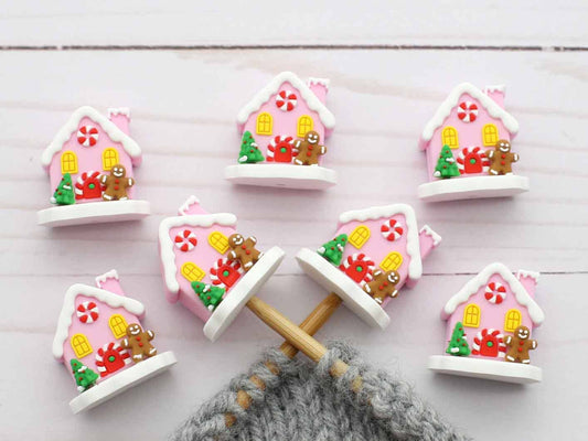 Six small decorative house-shaped stitch stoppers with pink roofs, white trim, candy motifs, and gingerbread figures, arranged on a white wood surface with knitting needles and gray yarn visible at the bottom.