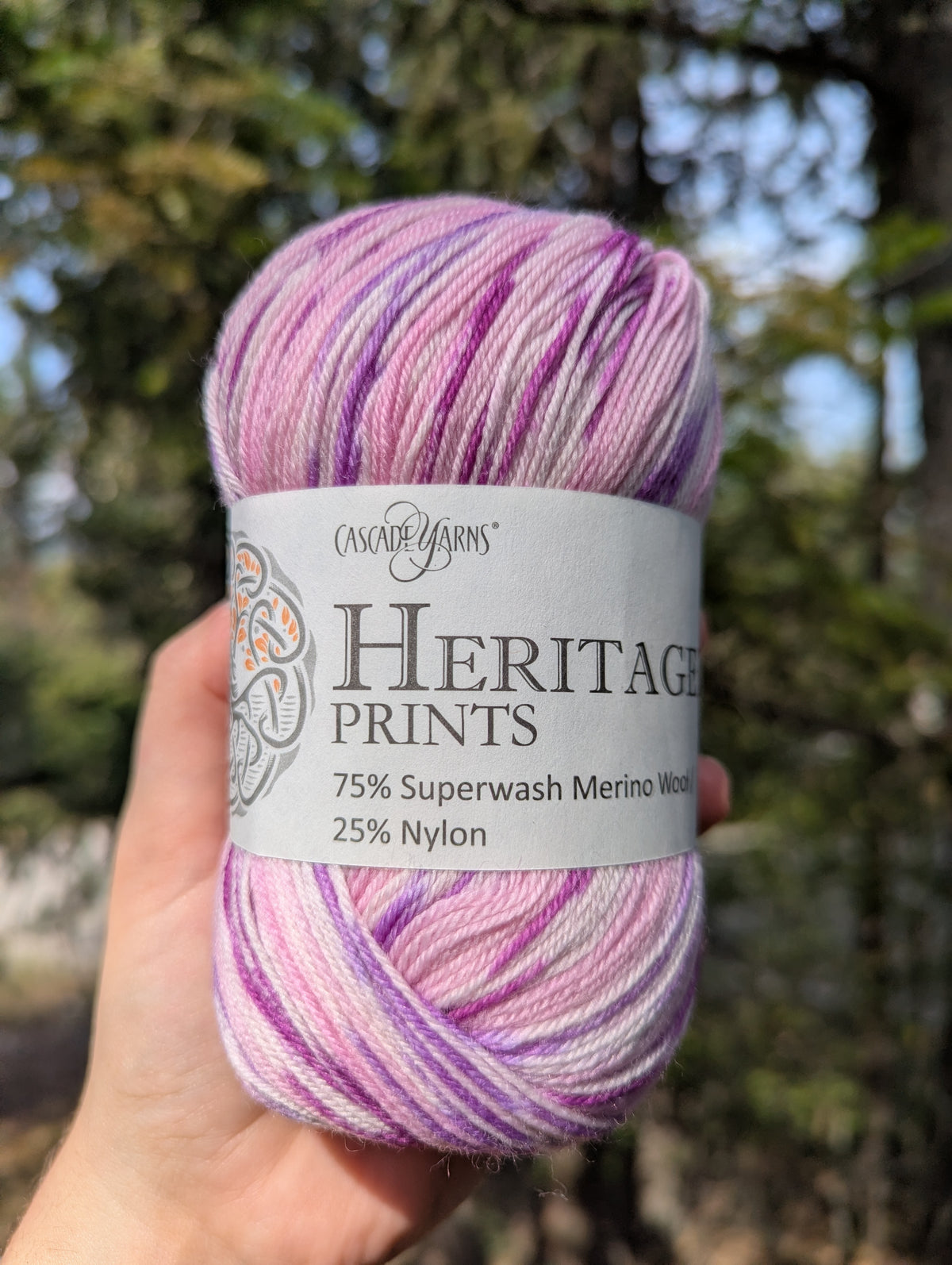 A hand holds a skein of Cascade Yarns Heritage Prints yarn, featuring a variegated pattern of purple, pink, and white merino wool with nylon. The background shows a blurred outdoor forest scene.