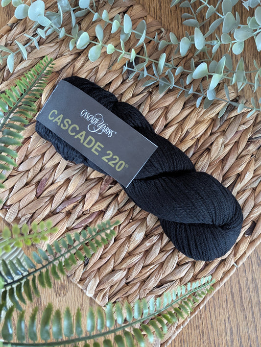 A skein of black Cascade 220 yarn wrapped with a black paper label, displayed on a woven straw mat with green foliage accents.