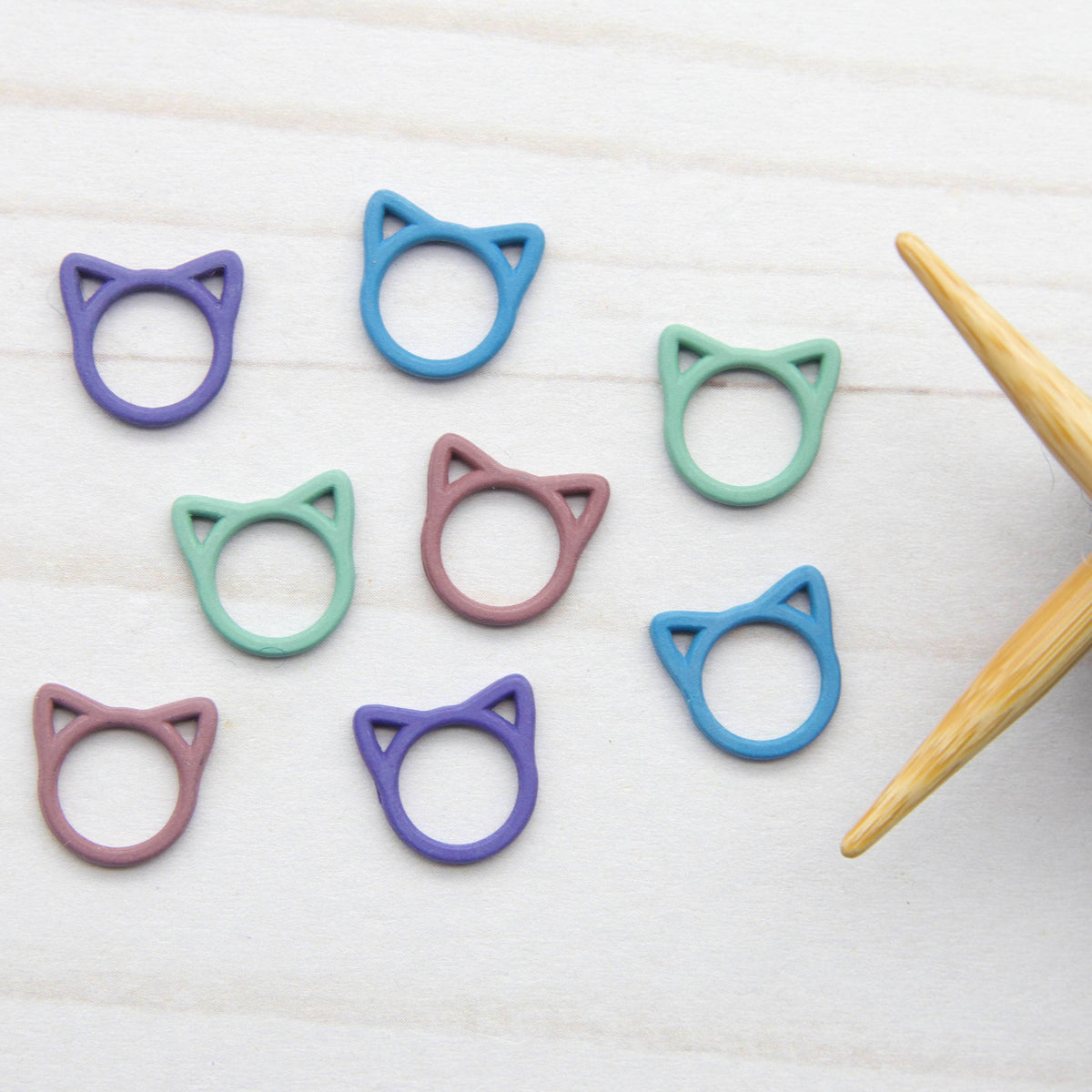 Colorful cat-shaped knitting stitch markers on a white surface with a wooden needle and gray yarn.