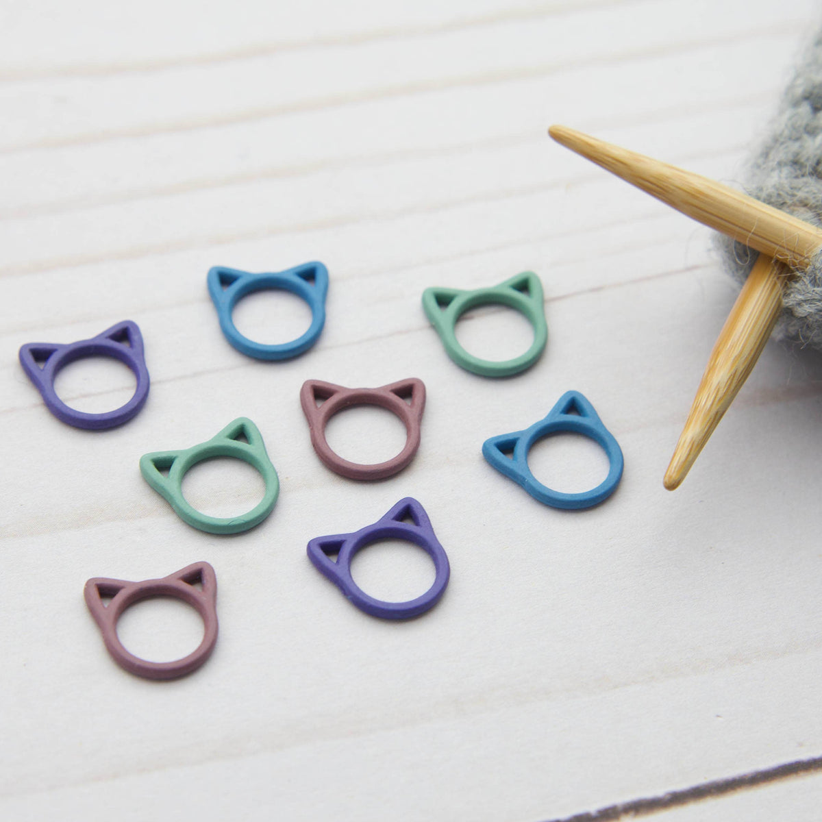 Colorful cat-shaped knitting stitch markers on a white surface with knitting needles.
