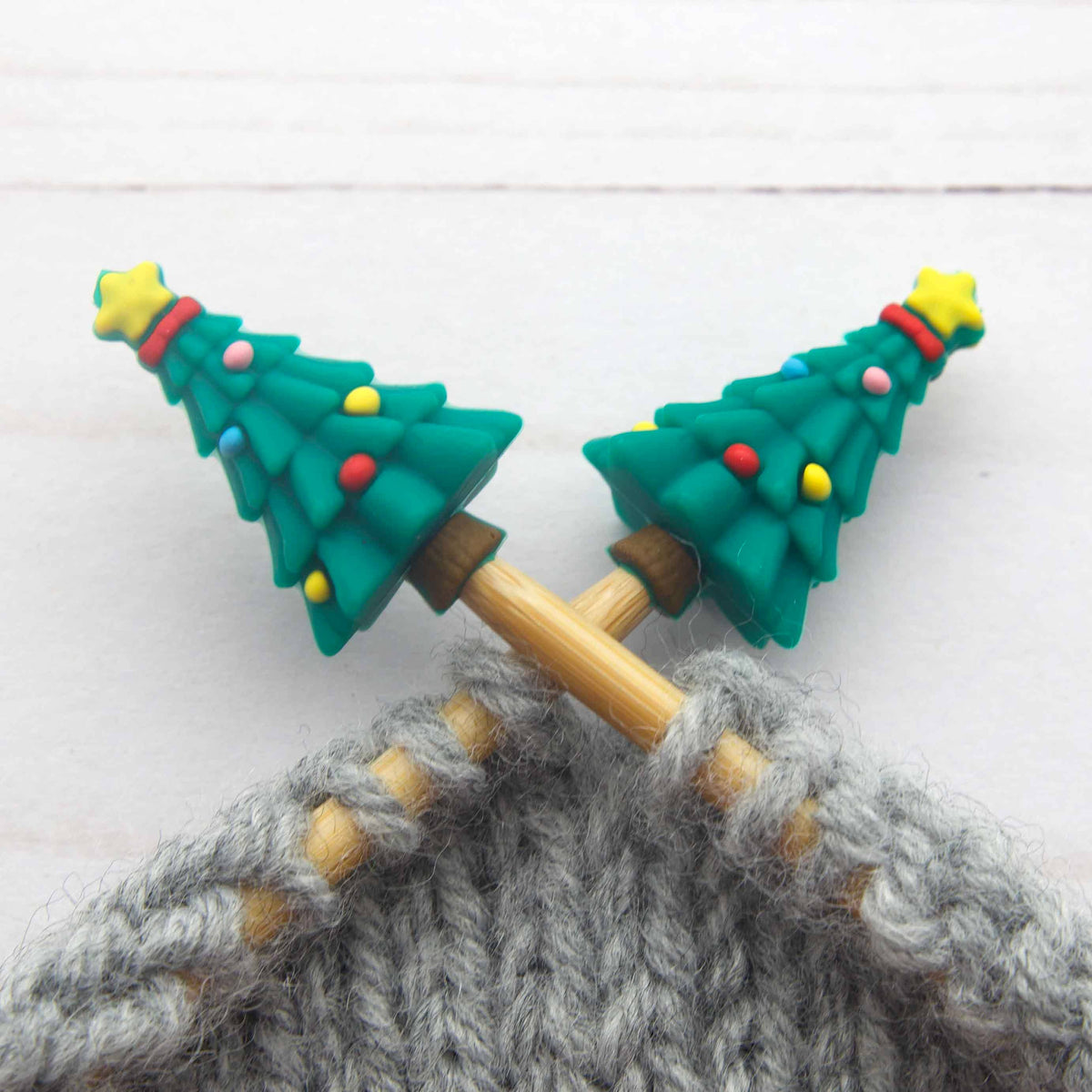 Close-up of two wooden knitting needles with green Christmas tree-shaped Stitch Stoppers decorated with small colorful ornaments, knitting gray yarn on a white wood background.