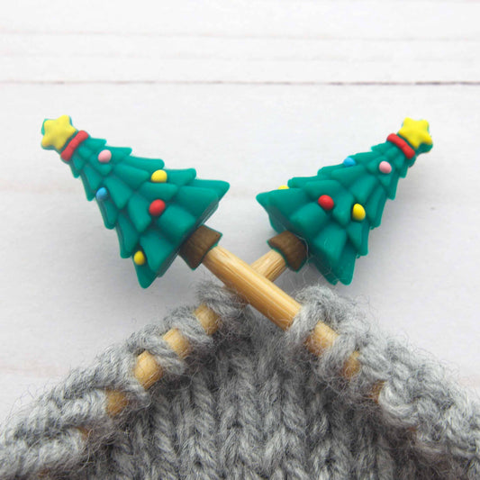 Close-up of two wooden knitting needles with green Christmas tree-shaped Stitch Stoppers decorated with small colorful ornaments, knitting gray yarn on a white wood background.