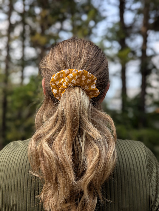 Person with long blonde hair tied back with a yellow scrunchie, standing outdoors with trees in the background.