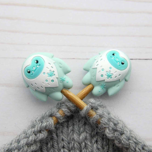 Two cute, cartoonish yeti stitch stoppers on knitting needles with mint green and white bodies, shown inserted into knitting needles with grey yarn on a light wood background.