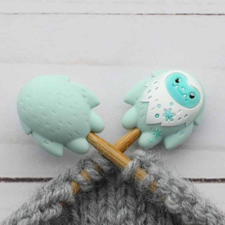 A close-up of two mint green and white yeti stitch stoppers on knitting needles with gray yarn, set against a light gray wood plank background.