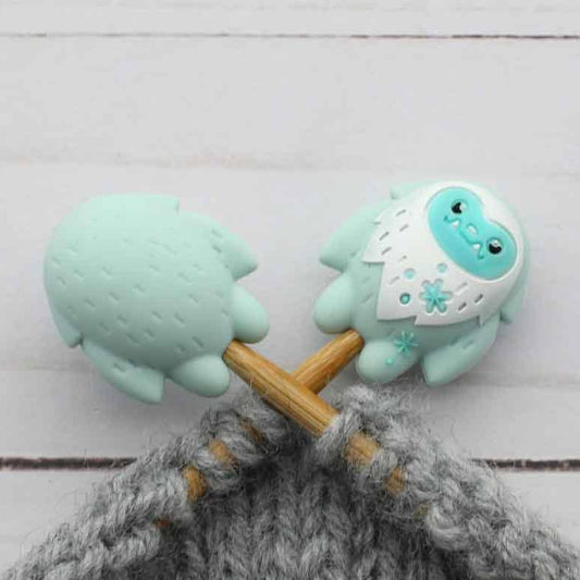 A close-up of two mint green and white yeti stitch stoppers on knitting needles with gray yarn, set against a light gray wood plank background.