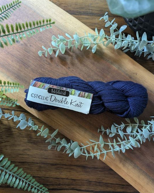 Navy skein of Estelle Double Knit yarn on a wooden board with eucalyptus leaves.