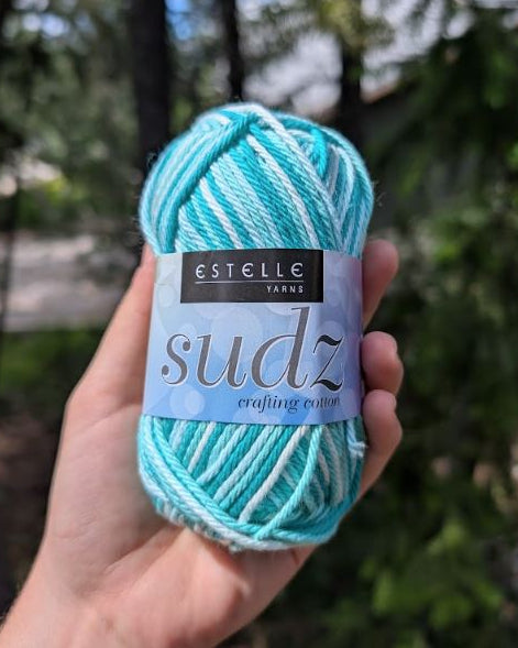 Turquoise yarn ball with Estelle Yarns label held in a hand against a blurred natural background