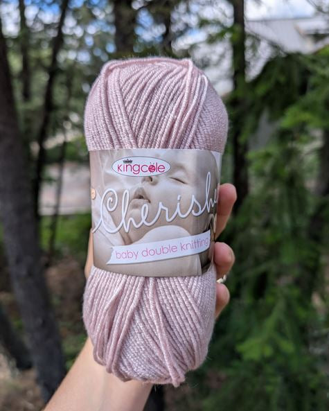 Ball of light purple yarn with 'King Cole Cherish' label held in front of a blurred natural background
