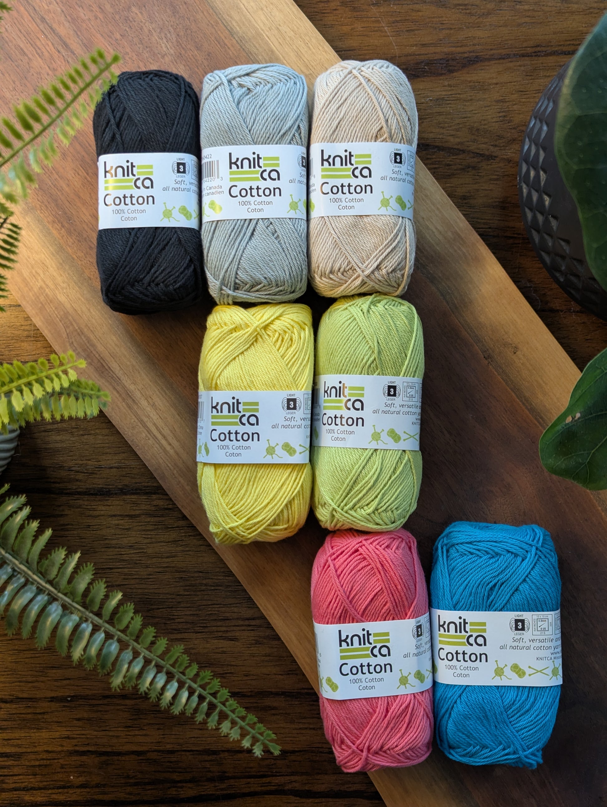 Six balls of knit cotton yarn in various colors on a wooden surface with plants around.