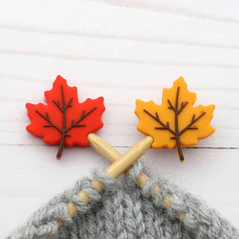 Two autumn maple leaf-shaped stitch stoppers in red and yellow on knitting needles on a white wood background.