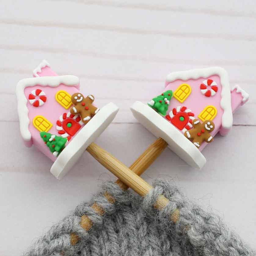 Two pink gingerbread house shaped stitch stoppers on knitting needles with wooden handles and gray yarn wrapped around them, placed on a light wood surface.