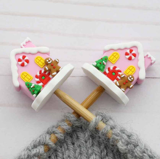 Two pink gingerbread house shaped stitch stoppers on knitting needles with wooden handles and gray yarn wrapped around them, placed on a light wood surface.