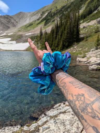 Hand holding a blue scrunchie with a mountain and lake background