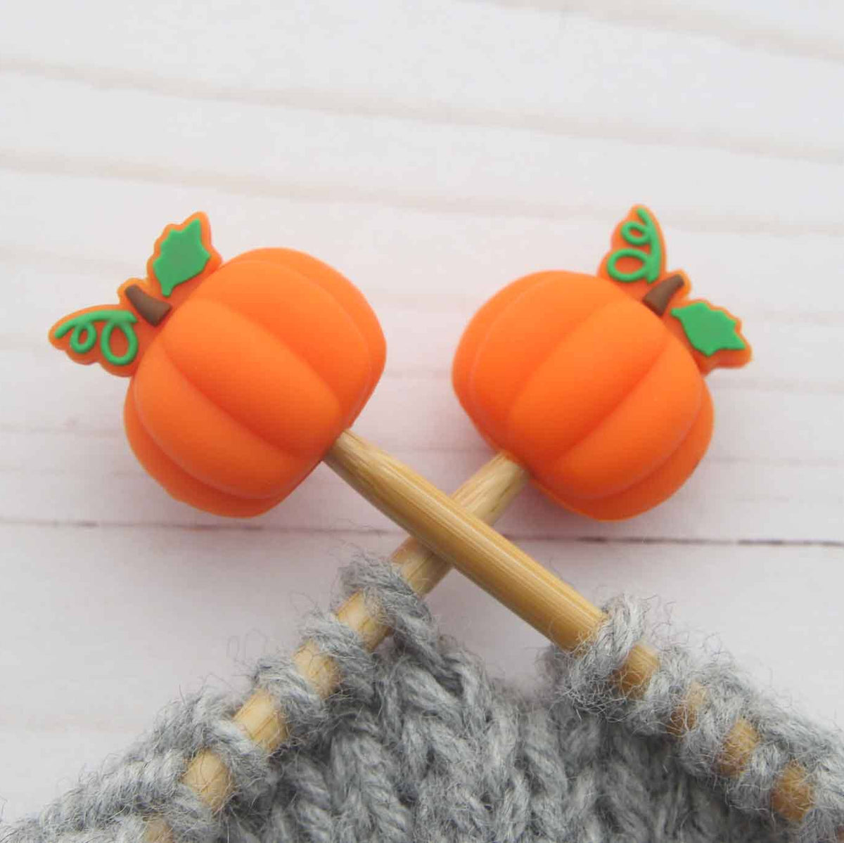 Bright orange pumpkin-shaped knitting Stitch stoppers with green leaf accents, shown inserted into knitting needles on a white wooden background.
