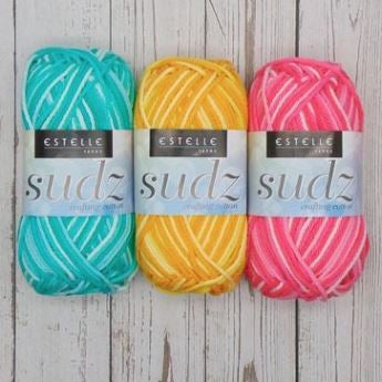 Three skeins of yarn in teal, yellow, and pink with 'Estelle' labels on a wooden surface.