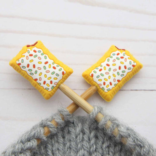 Two toaster pastries with sprinkles stitch stoppers on knitting needles