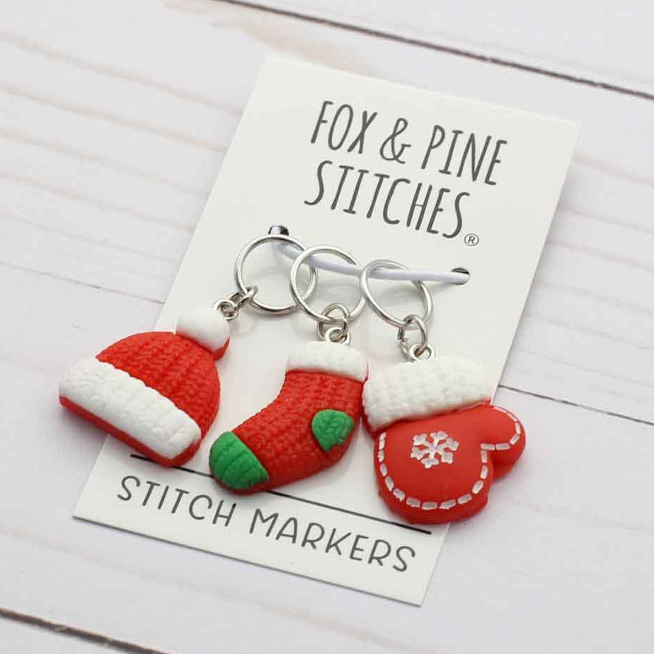 Three Christmas-themed stitch markers by Fox & Pine Stitches, featuring a Santa hat, a stocking, and a mitten, displayed on a white card over a white wood background.