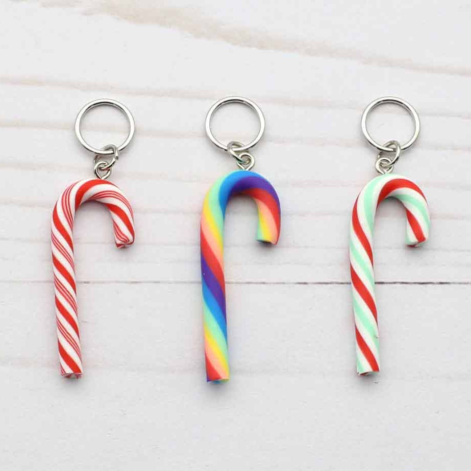 Three miniature candy cane stitch markers with silver metal keyrings, displayed on a light white wood background. The keychains feature three different designs: a classic red and white striped candy cane, a vibrant rainbow-striped candy cane, and a green, white, and red striped candy cane.
