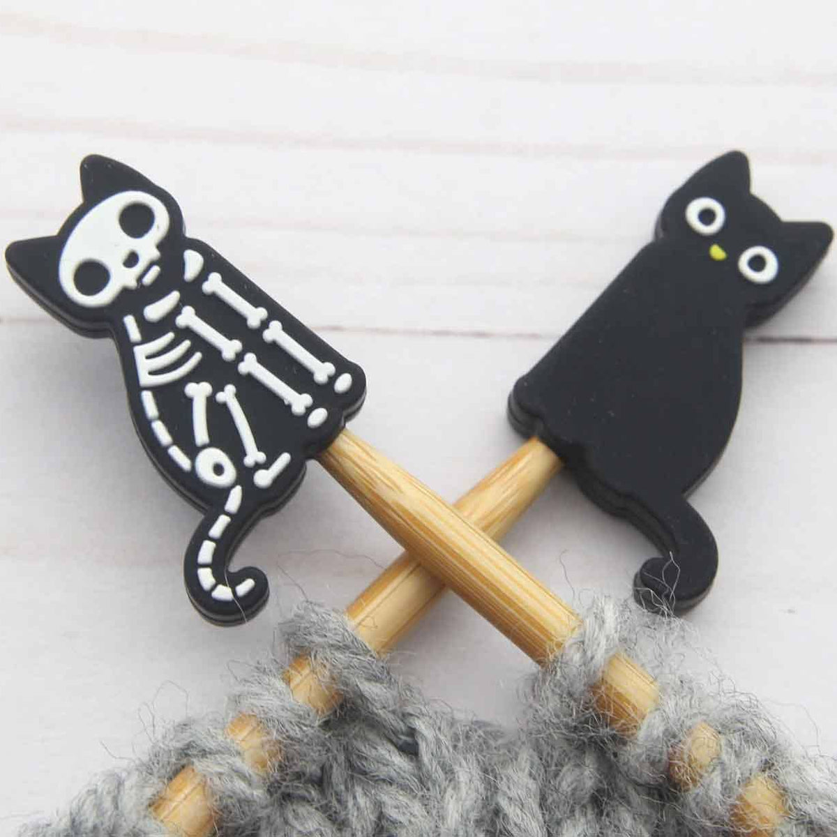 Two black cat-shaped stitch stoppers on knitting needles with white skeleton and solid cat designs, set against a light white wood background.