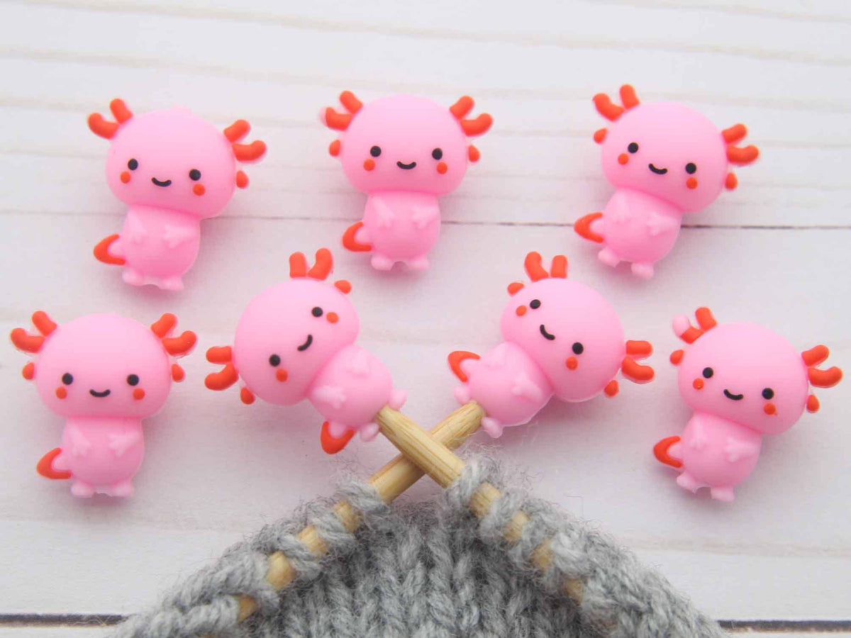 Pink axolotl stitch stoppers on a wooden surface with a gray knitted fabric.