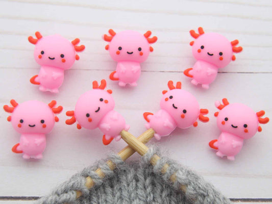 Pink axolotl stitch stoppers on a wooden surface with a gray knitted fabric.
